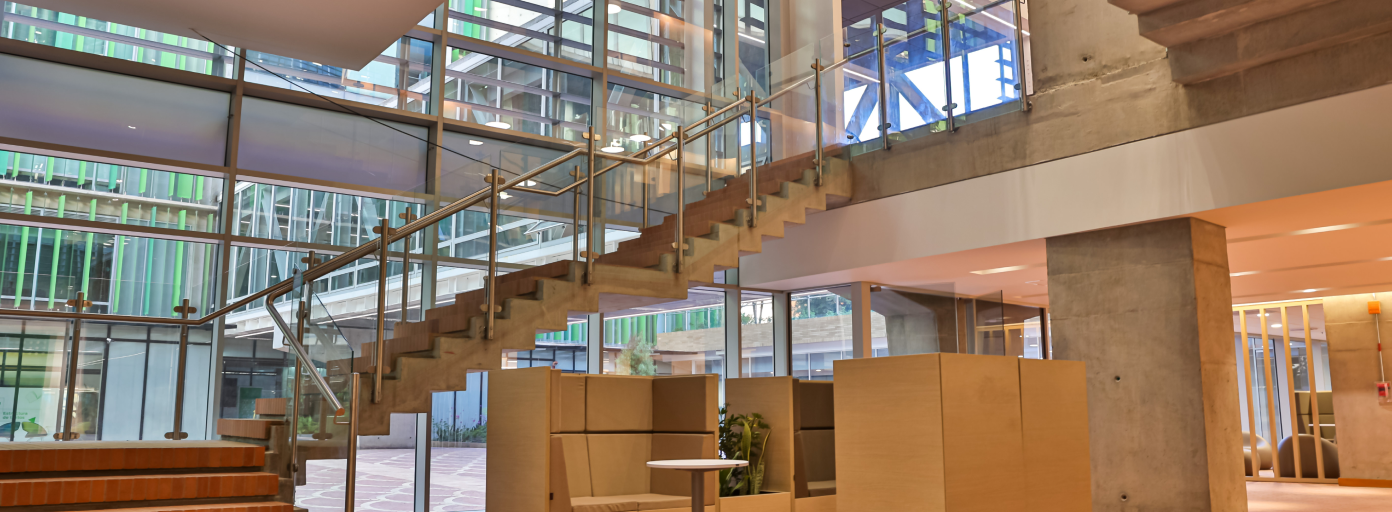 A modern interior featuring a spacious area with a large glass facade. A concrete staircase with a glass railing ascends to the upper level. The room is filled with natural light and has wooden furniture and partitions.